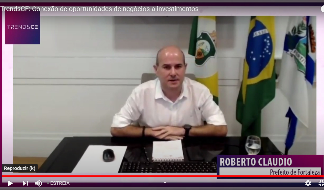 print de vídeo com o prefeito sentado à mesa e bandeiras ao fundo